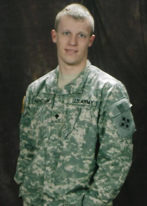 A person in a U.S. Army uniform stands in front of a dark background, looking at the camera with a slight smile. The uniform features a name tag and a shoulder patch.