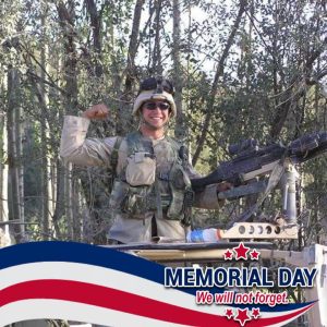 A smiling soldier in uniform flexes his arm while standing next to a mounted gun in a wooded area. The image features a Memorial Day: We will not forget banner with red, white, and blue stripes at the bottom.