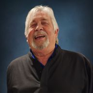 An older man with white hair and a beard is smiling and laughing, wearing a dark shirt, against a blue textured background.